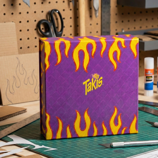 Prototipo de caja rígida para Takis con diseño de llamas moradas y amarillas sobre una mesa de trabajo en un taller de diseño.